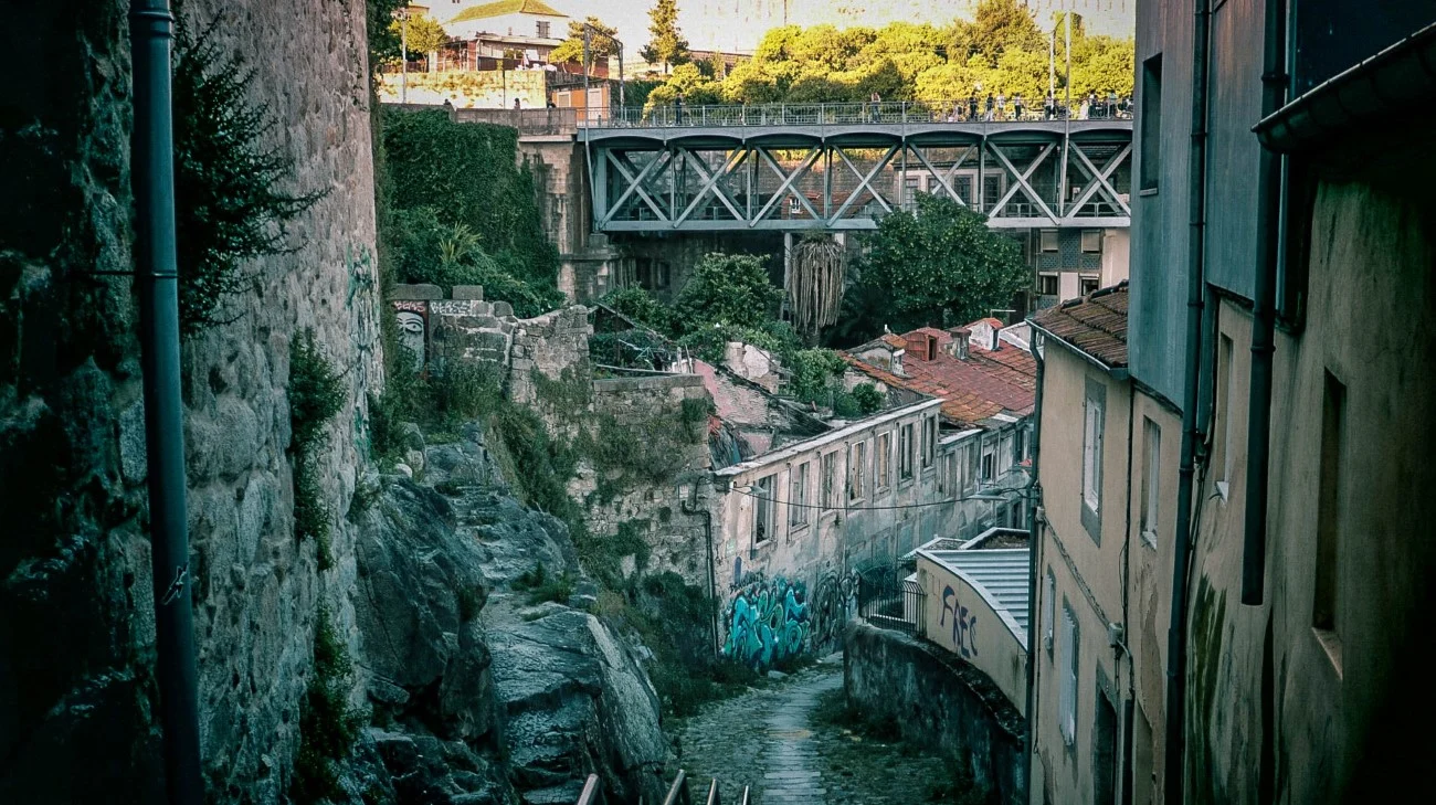 A metal footbridge crossing over a deep ravine with old buildings clinging to the cliffside