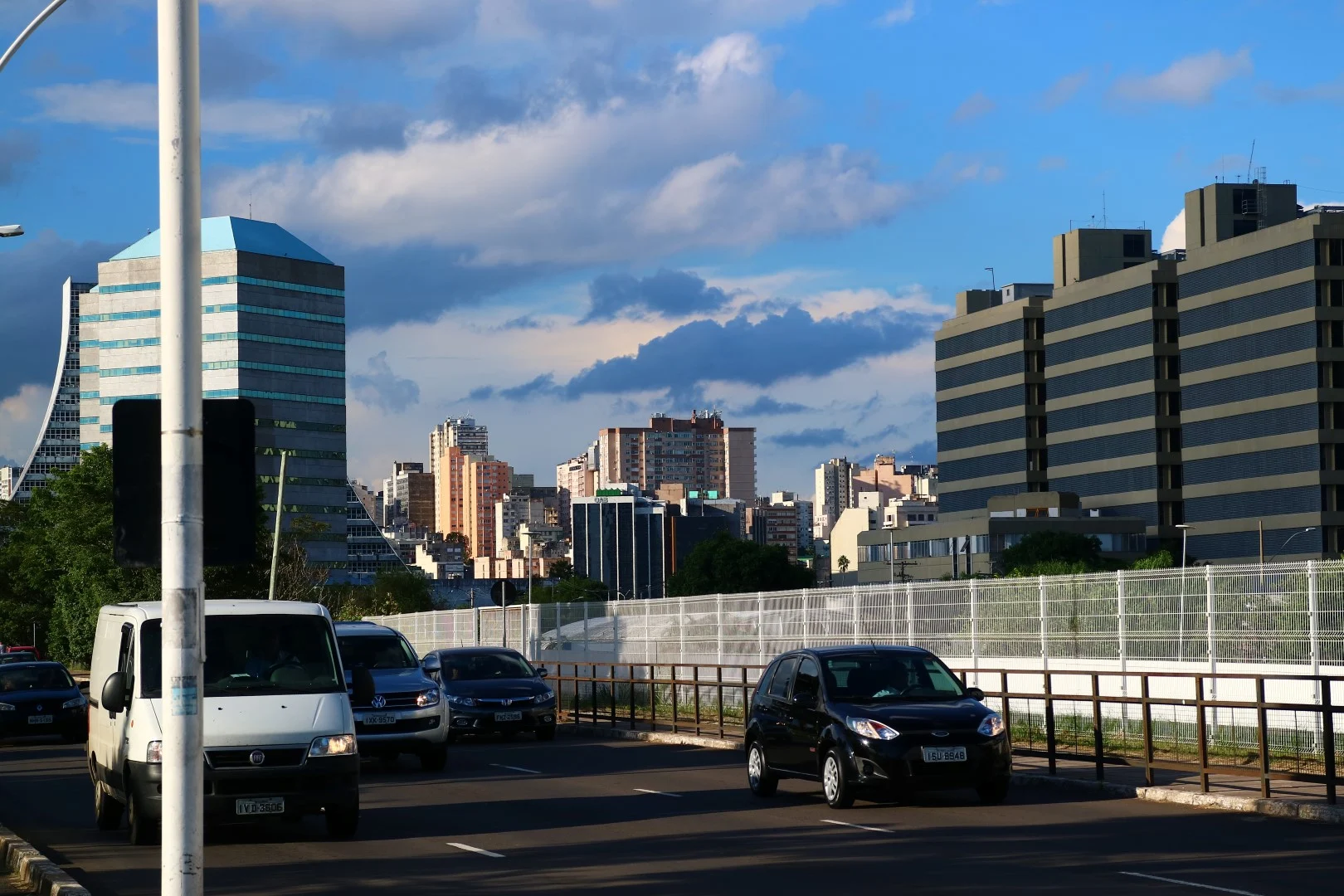 Downtown towers and an office block in cyan glass behind a wide boulevard, shot from a flyover ramp.