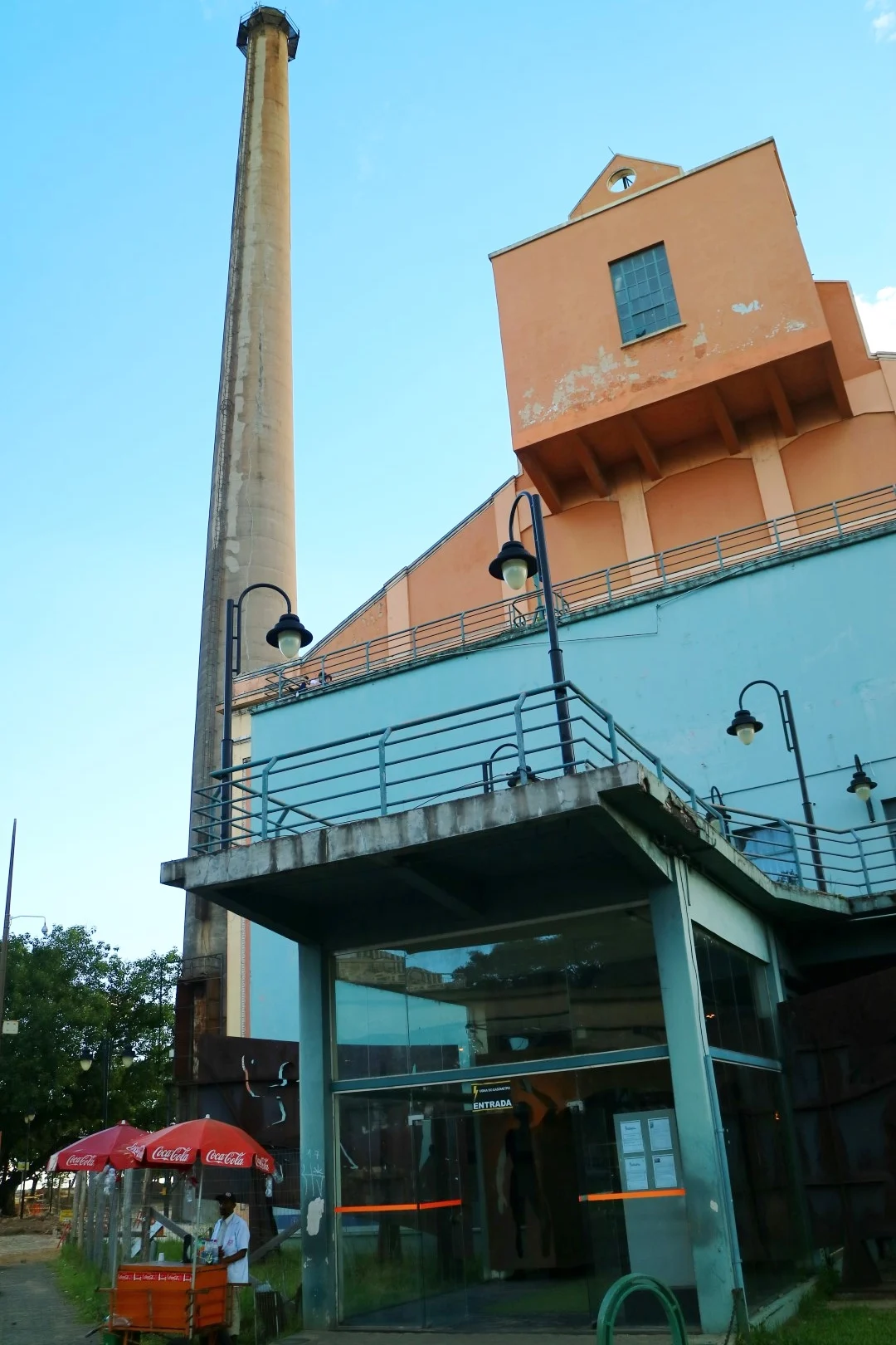 The Gasômetro from its entrance side: the salmon and turquoise brickwork, the connected utility block, a Coca-Cola umbrella vendor waiting outside.